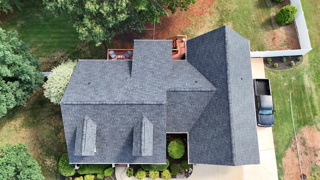 Aerial view of a house with a dark gray roof, surrounded by trees and a driveway with a parked car.