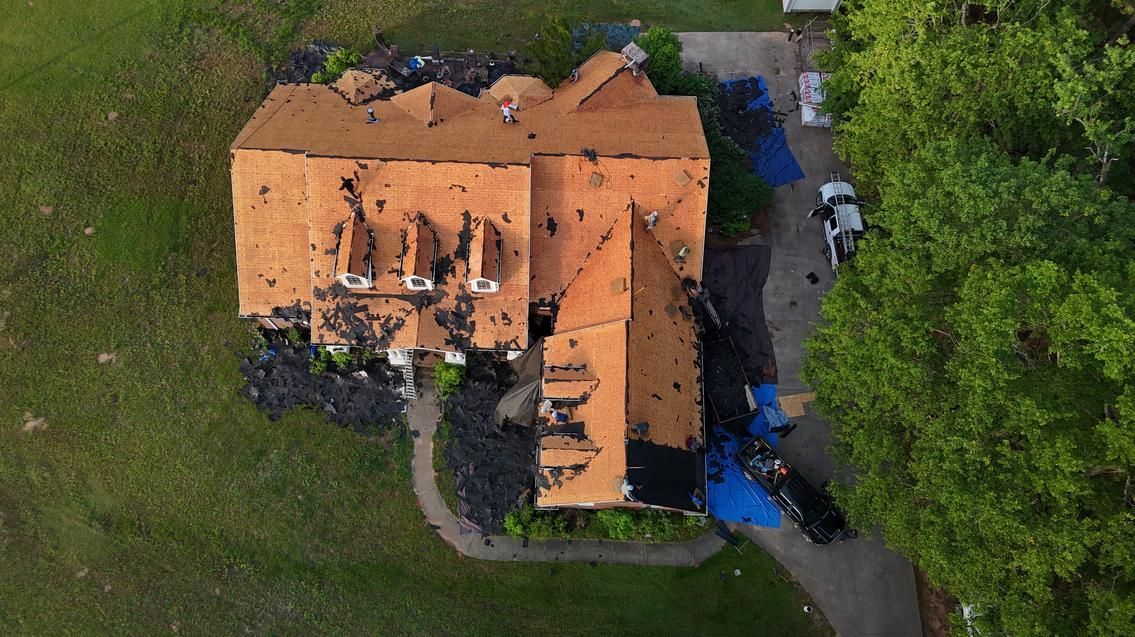Aerial view of a house with an orange roof surrounded by green trees and grass, with a driveway beside it