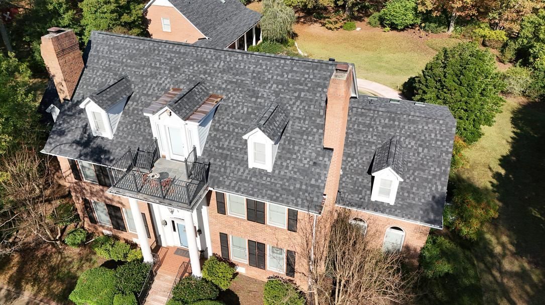 Aerial view of a large brick house with a dark roof, white dormers, and a tree-lined yard.