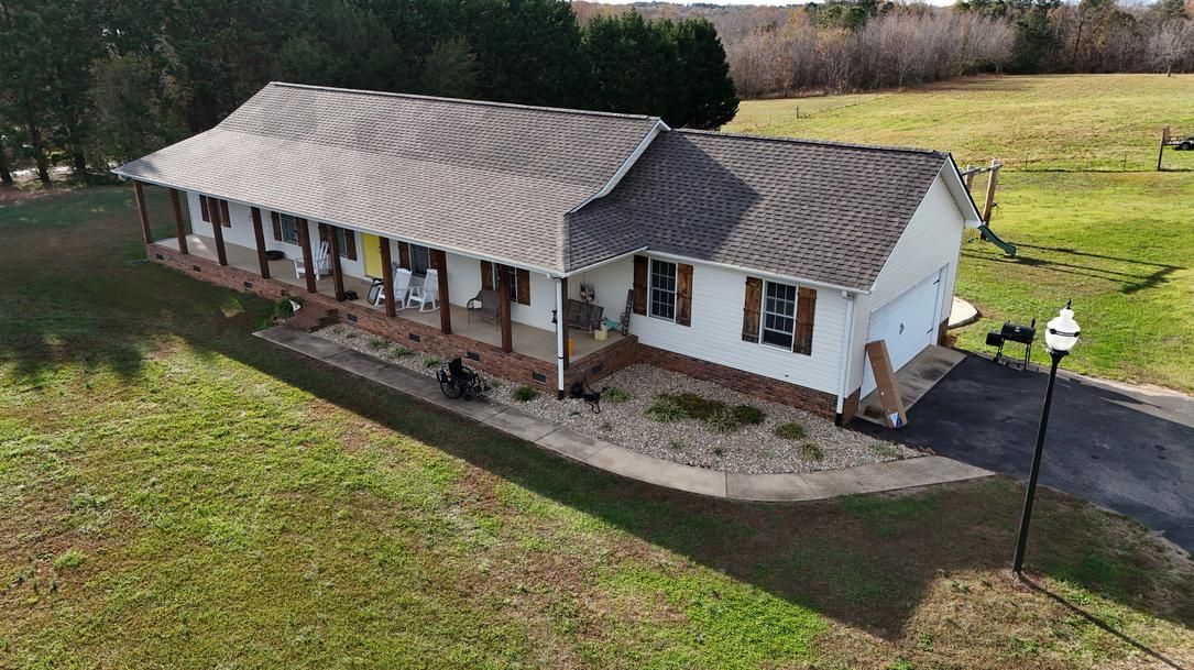 Aerial view of a single-story white house with a gray roof beside a grassy yard and driveway.
