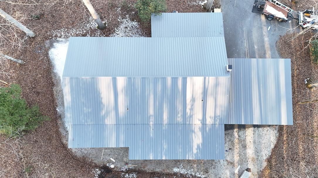 Aerial view of a small corrugated metal roof with an attached covered porch, surrounded by trees.