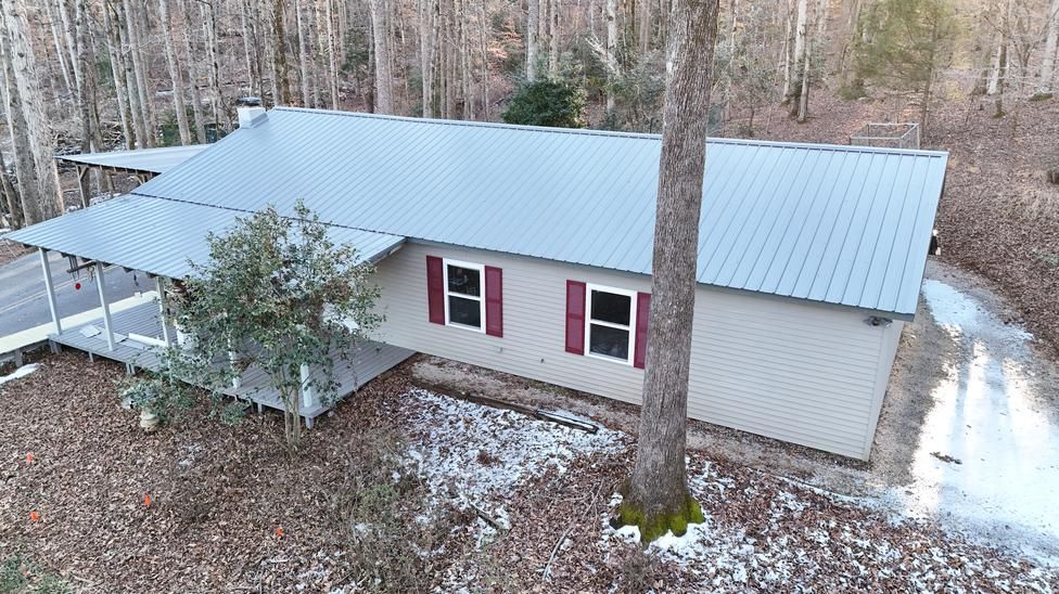Aerial view of a gray metal-roofed house with red shutters, surrounded by woods in late fall.