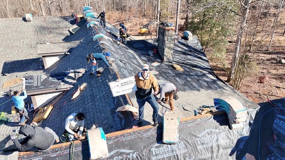 Workers installing shingles on a steep roof at a construction site, with tools and materials spread out.