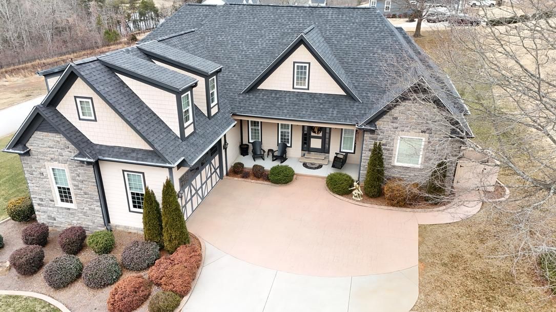 Aerial view of a large stone house with dark roof and curved driveway in a suburban setting