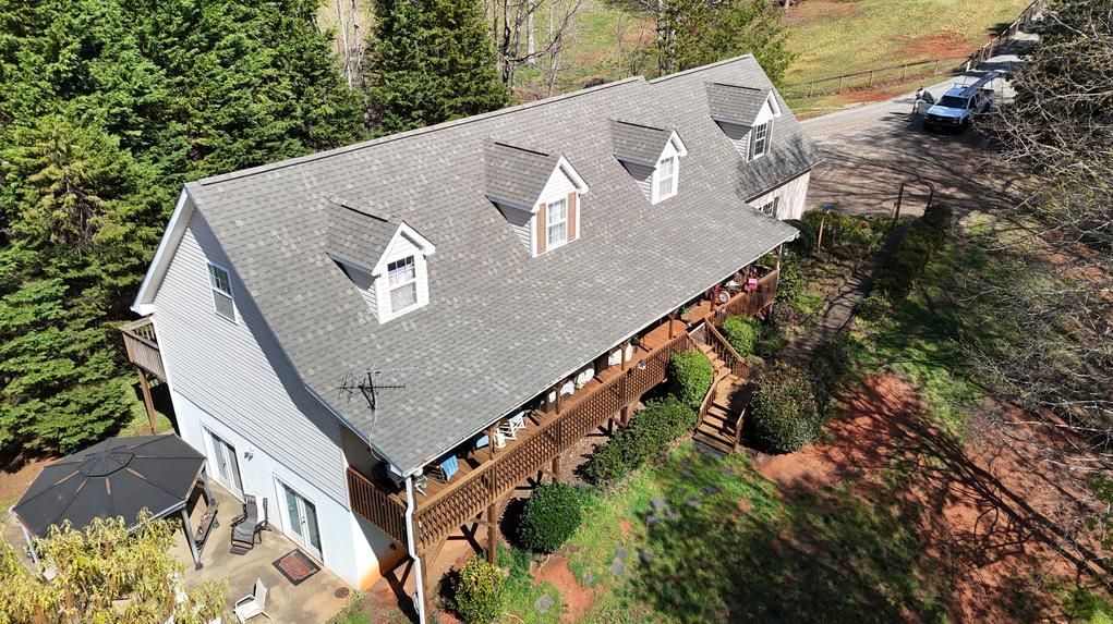 Aerial view of a white house with a gray roof, dormer windows, and a covered porch surrounded by trees.