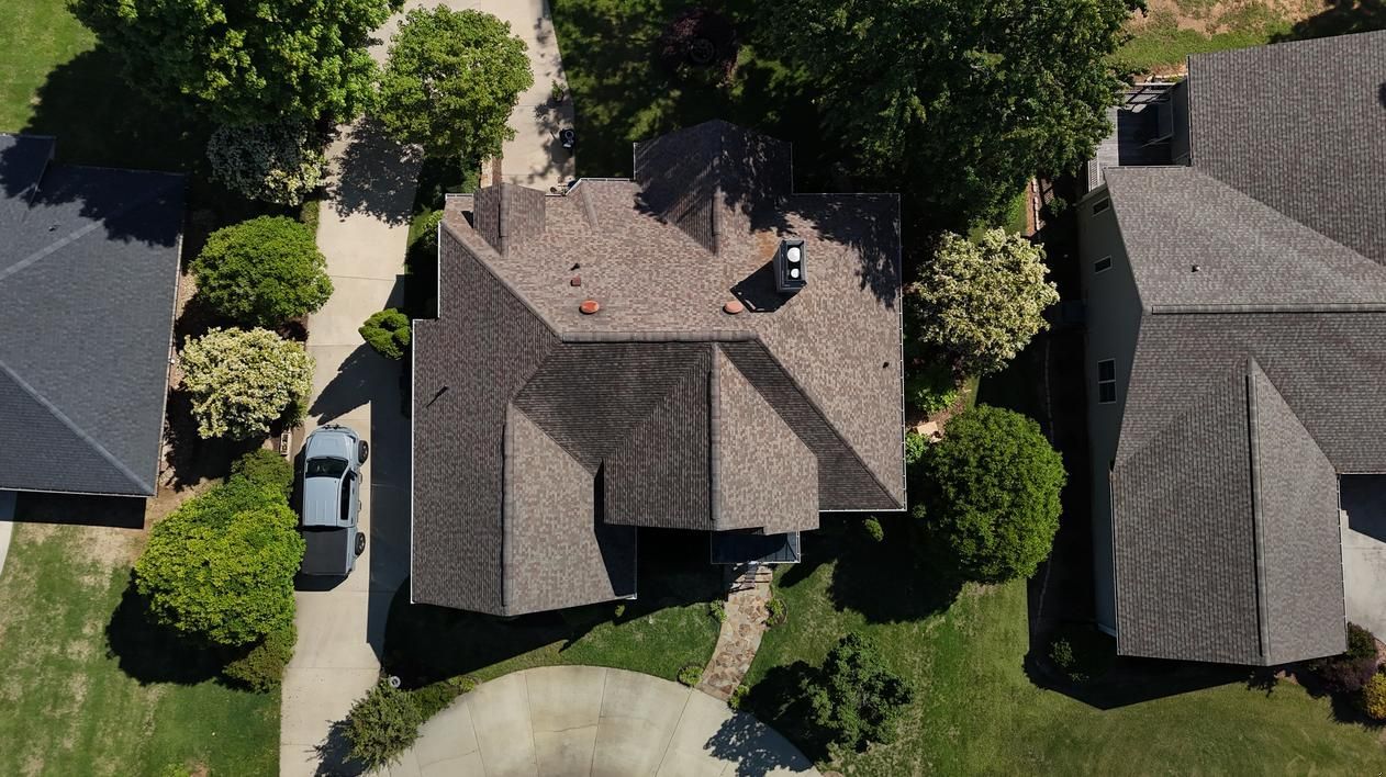 Aerial view of three suburban houses with gray roofs, trees, driveways, and parked cars.