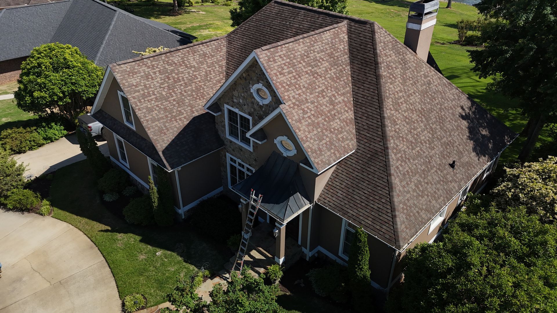 Aerial view of a large brown-roofed suburban house with a driveway and surrounding trees.
