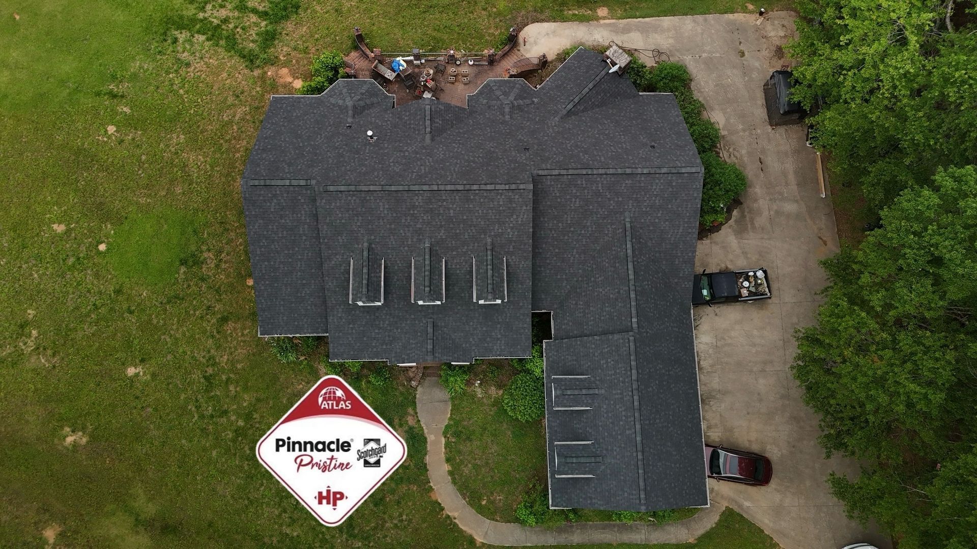 Aerial view of a dark-roofed house with a driveway, lawn, and a driveway sign near the front yard.