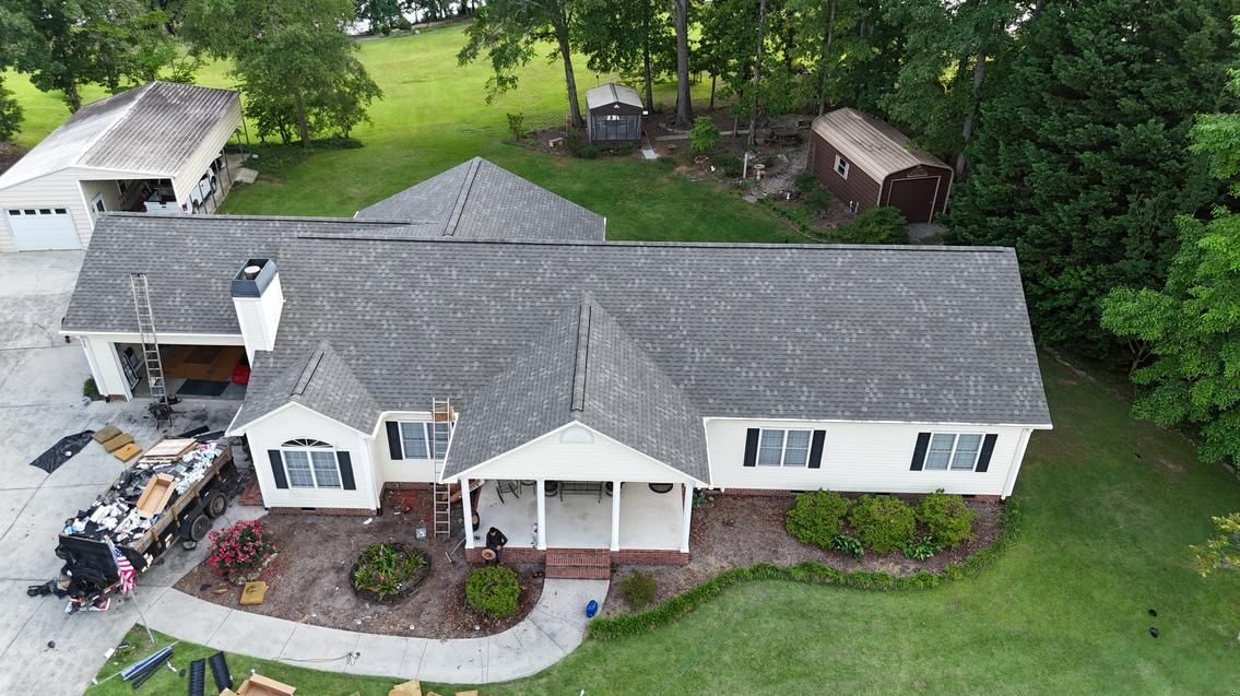 Aerial view of a white house with gray roof, front porch, and landscaped yard surrounded by trees.