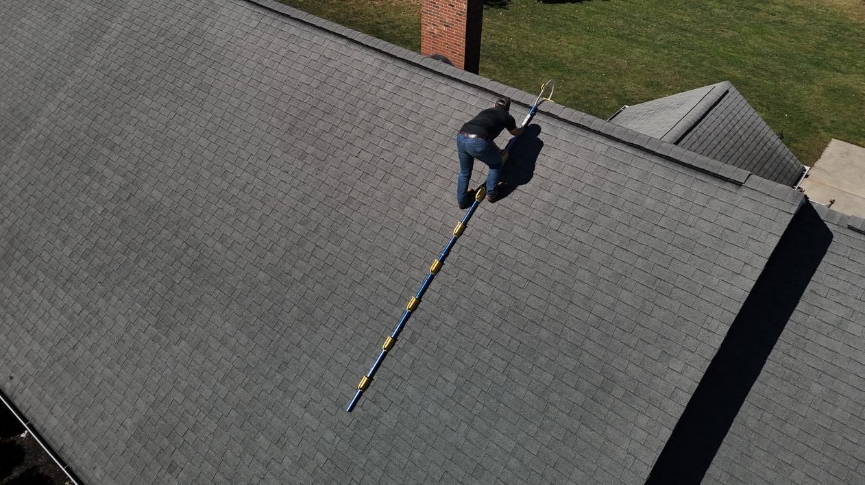 Person working on a gray shingled roof with a long extension ladder nearby