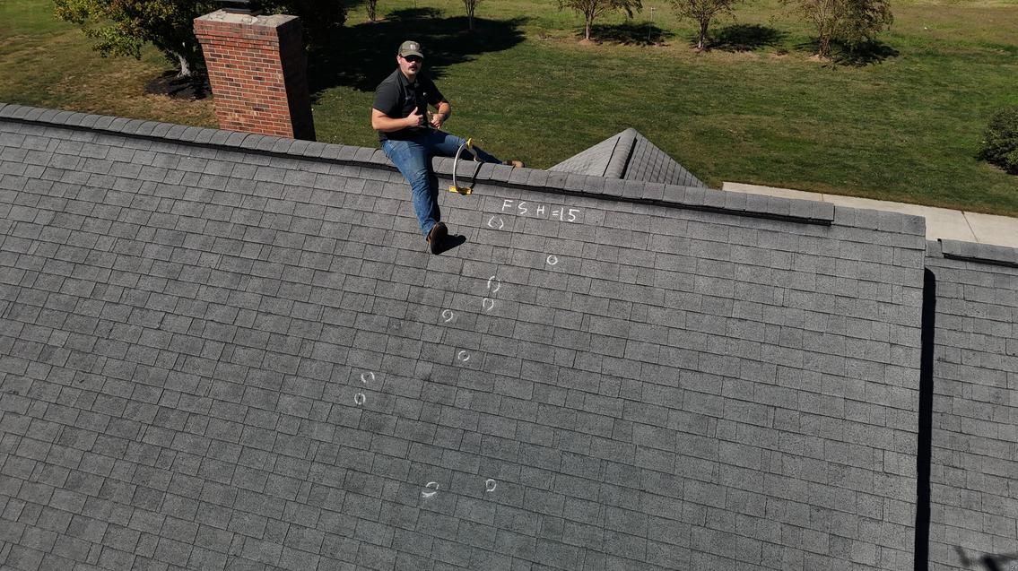 Person standing on a gray shingled roof near a brick chimney, holding a tool.