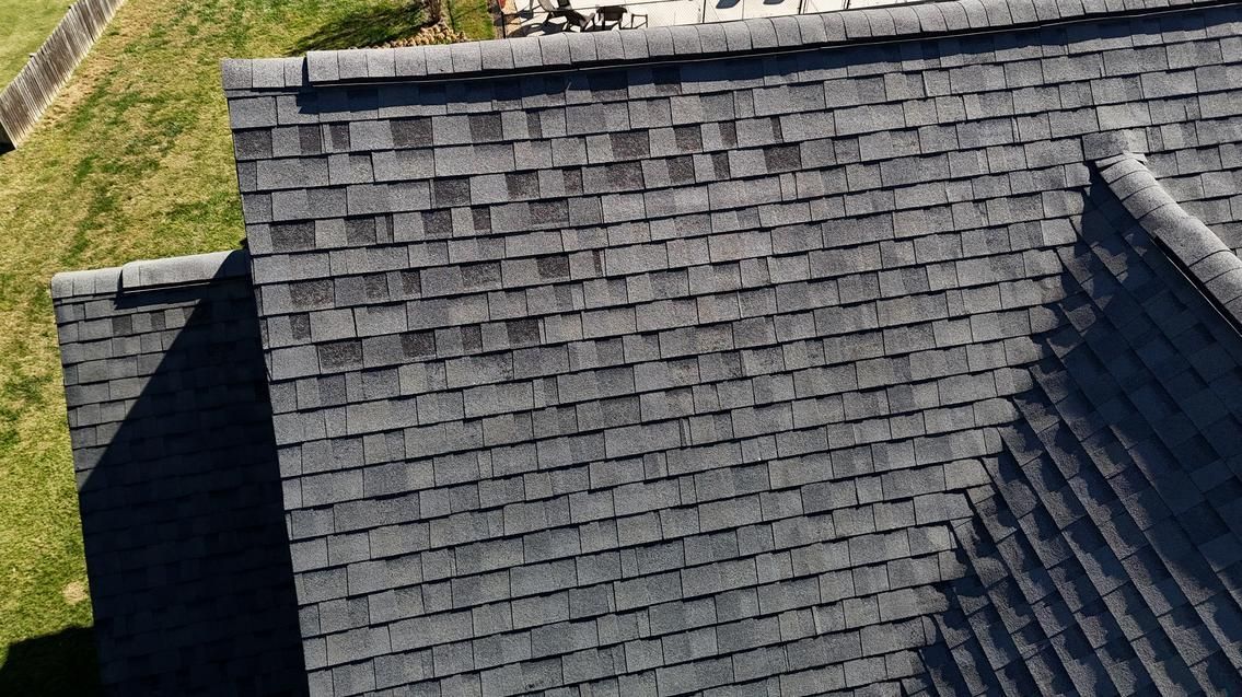 Aerial view of a dark gray shingled roof with dormer windows and a grassy yard edge