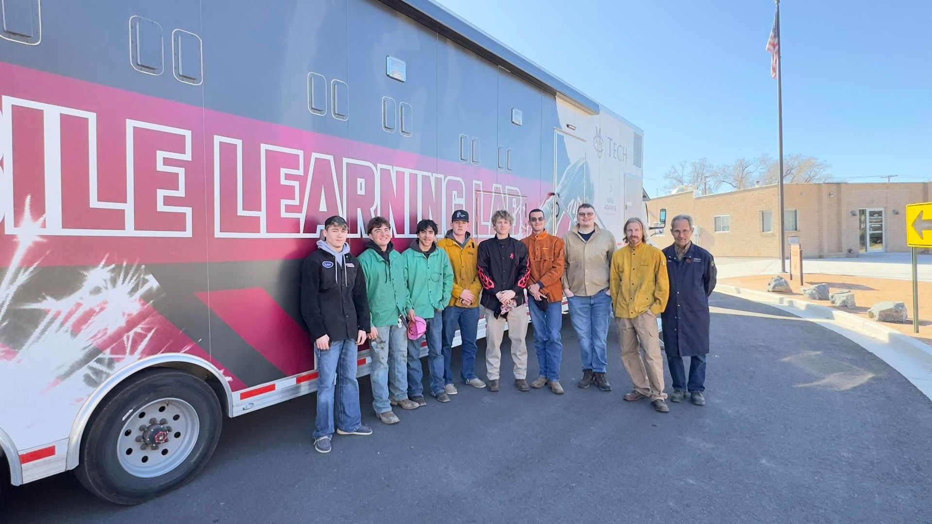 Group of people posing beside a red-and-blue “Little Lear…” vehicle on a sunny street.