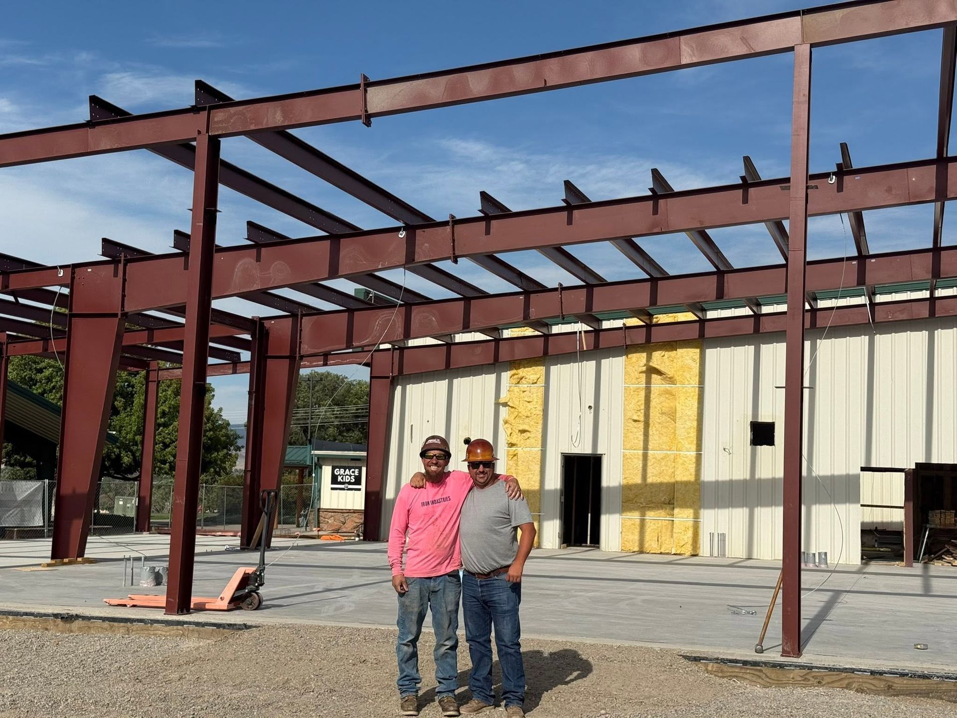 Two people standing in front of a steel-frame building under construction, with a yellow wall in the background.