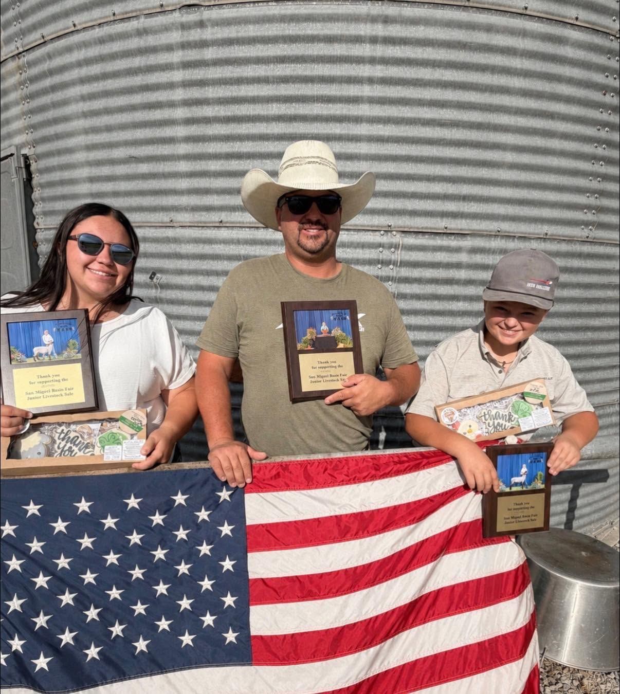 Three people holding plaques pose behind a draped American flag in front of a metal silo.
