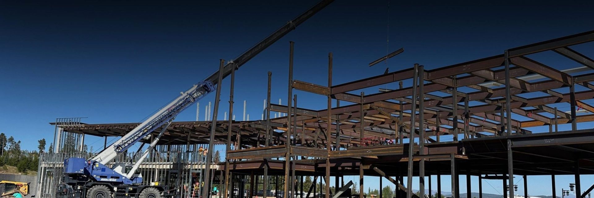 Steel-frame building under construction with a crane and workers on a sunny site