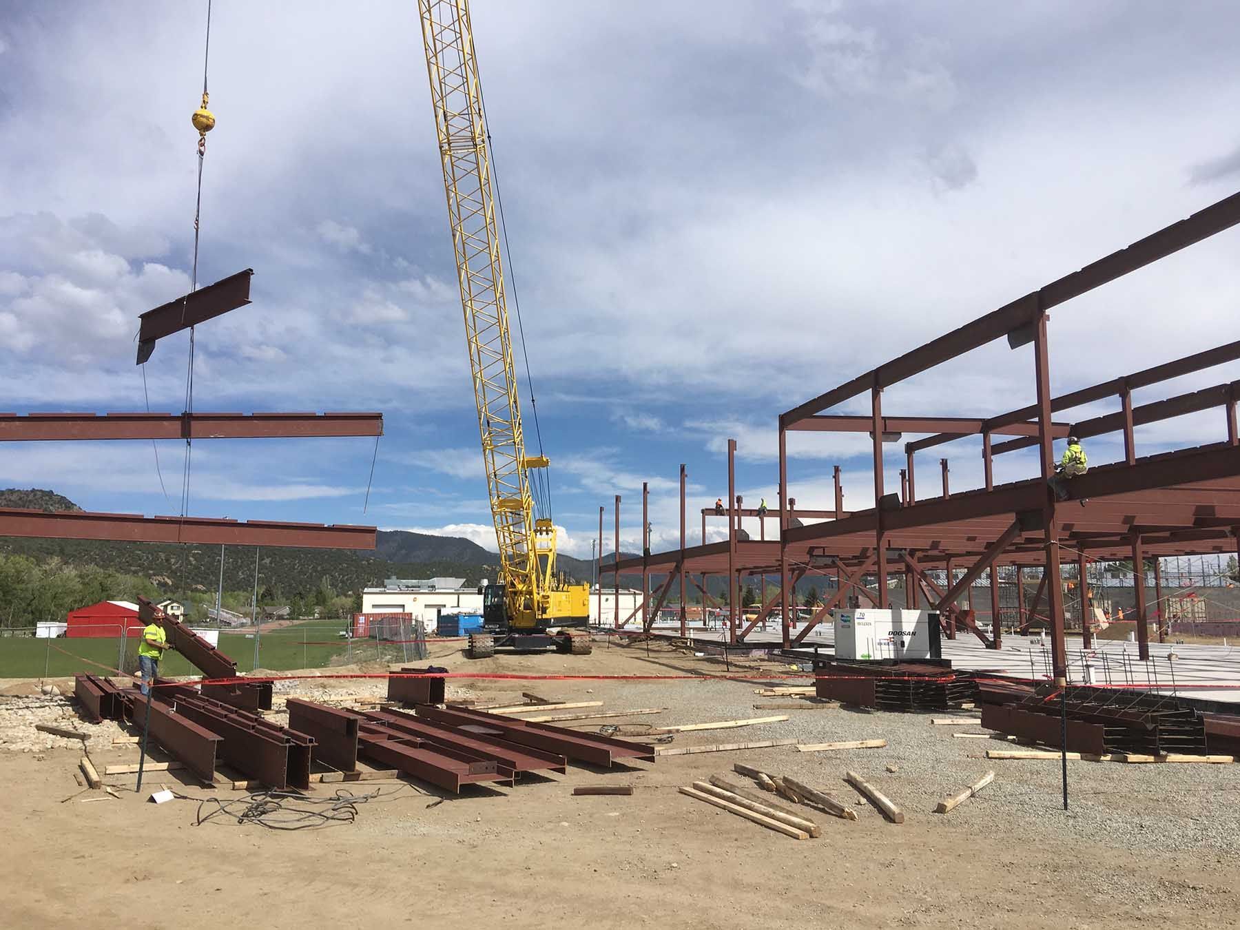 Construction site with crane lifting steel beams beside a partially built metal structure under cloudy sky