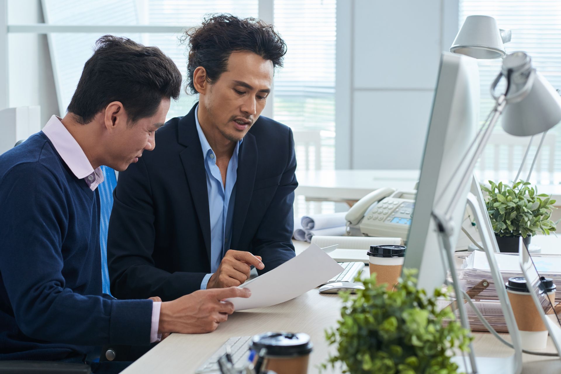 Two men in an office looking at a document. One wears a blazer, the other a sweater. Desk with a computer and plants.