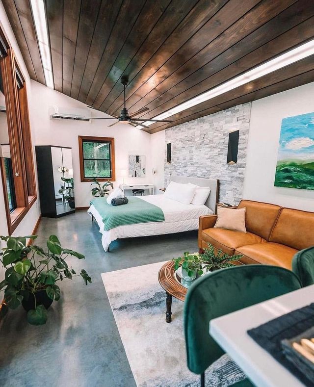 Modern bedroom with wood ceiling, white bed, tan sofa, plants, and soft natural light