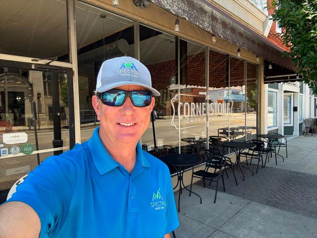 Man in blue polo and cap taking a selfie outside a cafe with patio seating and string lights