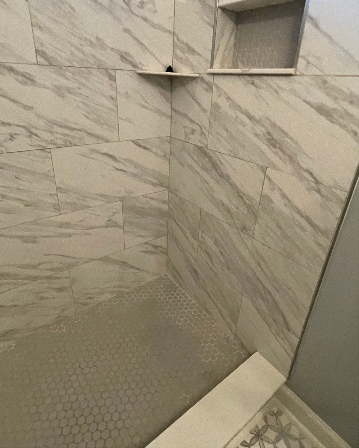 Tiled shower with marble-look walls and hexagonal floor. Built-in shelf and bench. Light gray tones.