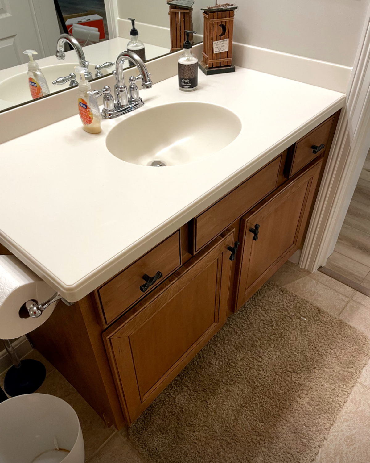 Bathroom vanity with a white countertop, oval sink, and wooden cabinets. There is a mirror, soap dispenser, and a rug.