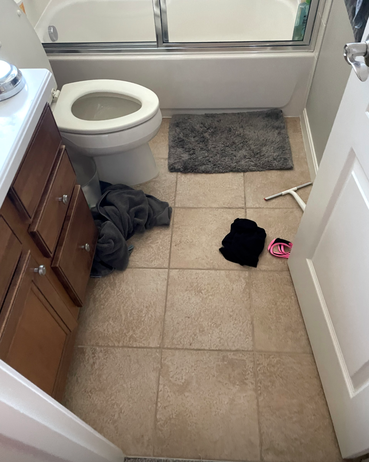 Bathroom interior: toilet, vanity, bath/shower, towels on floor, rug, open door.
