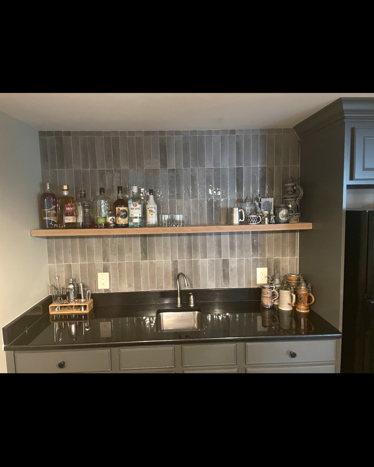 Home bar with wood shelf, liquor bottles, sink, black countertop, gray cabinets, and a gray-tiled backsplash.