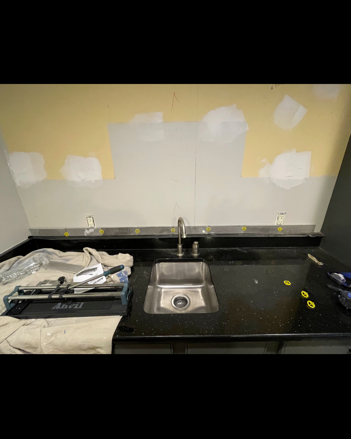 A kitchen counter with a sink, faucet, and tiling tools. Yellow and gray wall with patching.