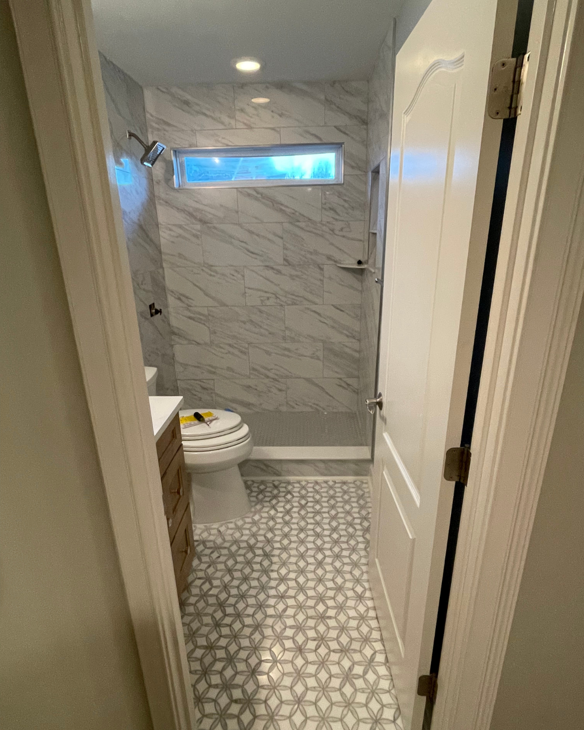 Bathroom interior: white marble shower, patterned floor, toilet, vanity, and closed white door.