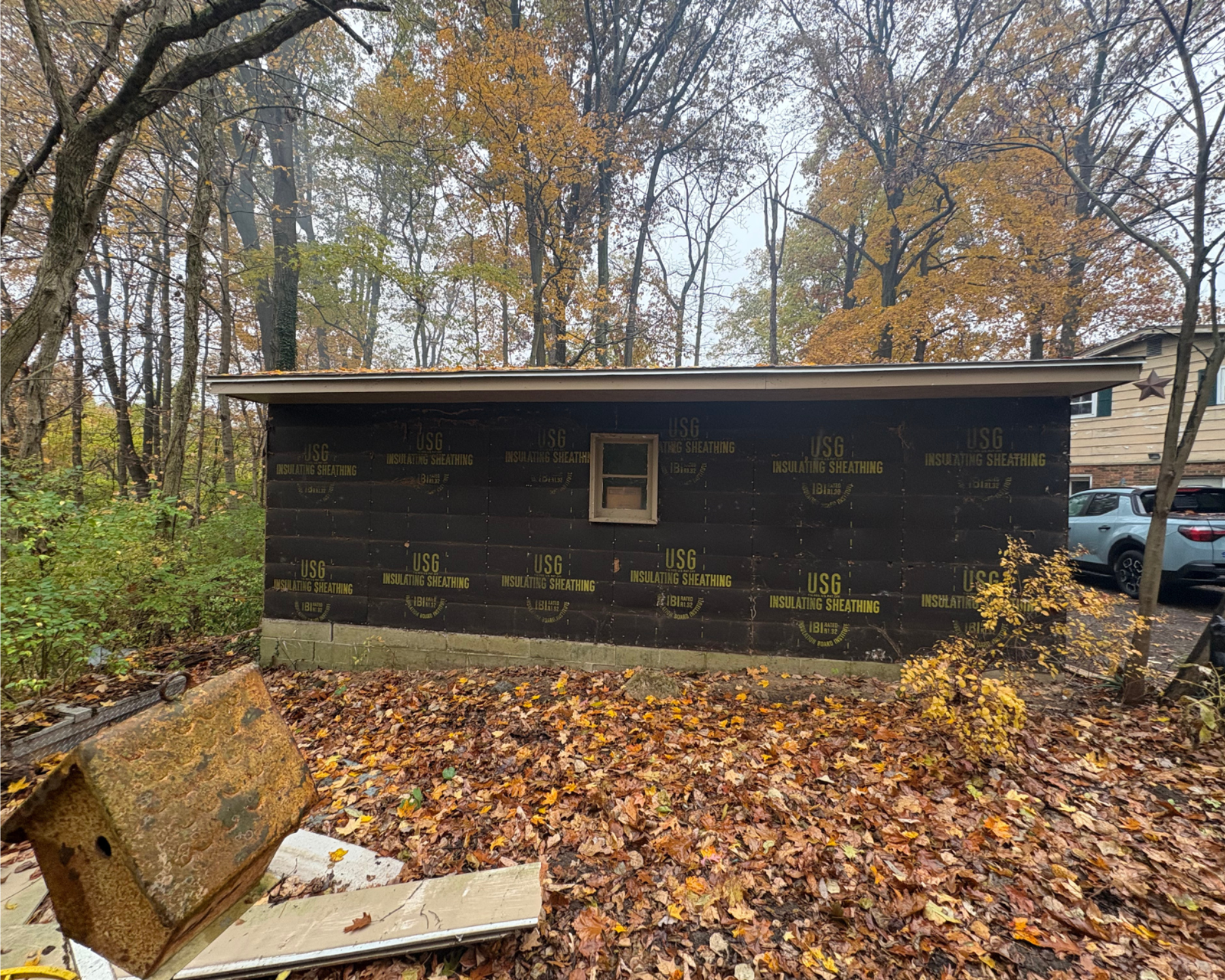 A shed under construction in a wooded area; black wrap on the walls, small window, brown roof.