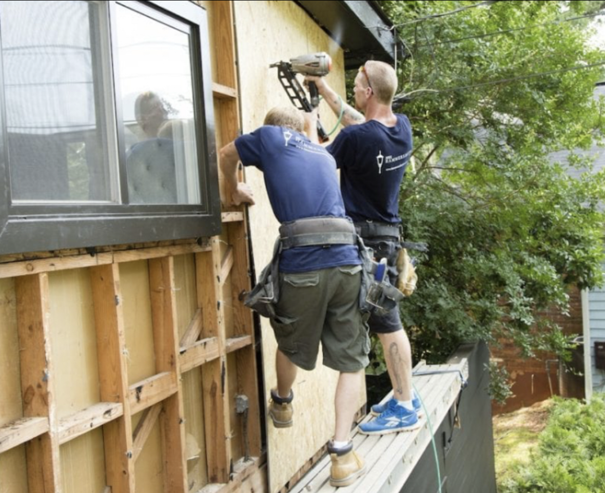Two people installing siding on a building's exterior near a window, one using a nail gun.