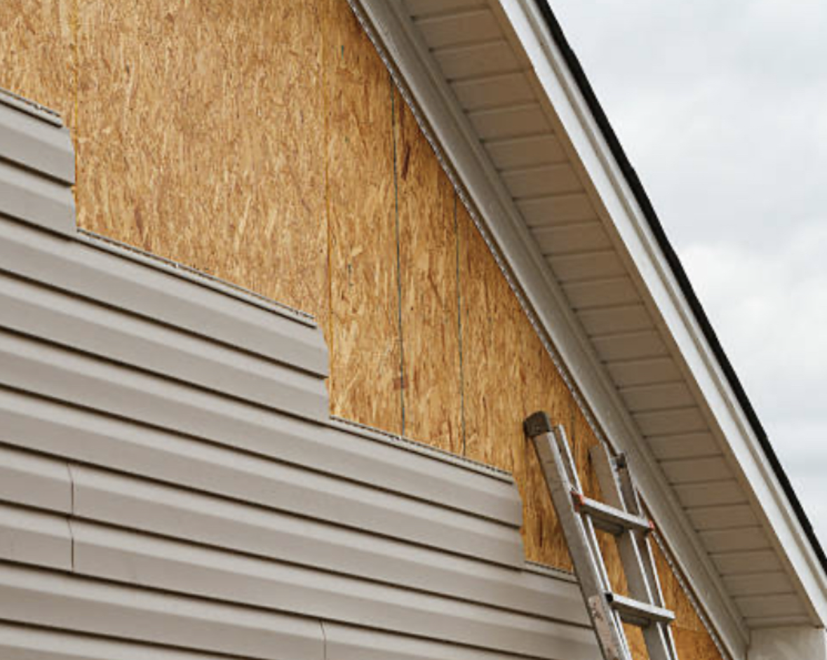 House siding partially installed; ladder leaning against the wall. Brown particle board exposed.