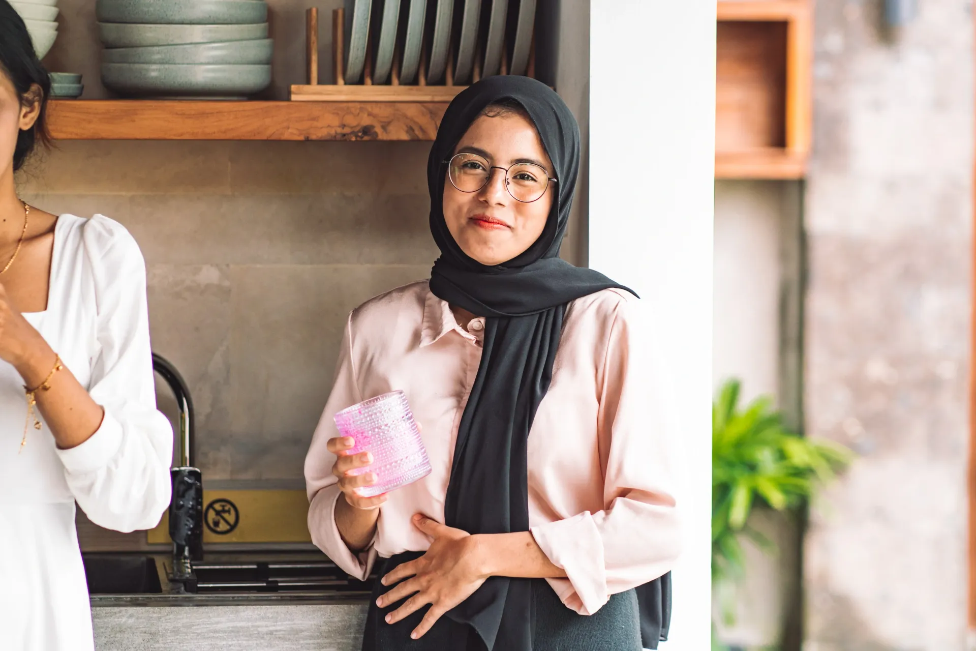 Wanita berhijab memegang barang berwarna merah muda, tersenyum, di dapur.