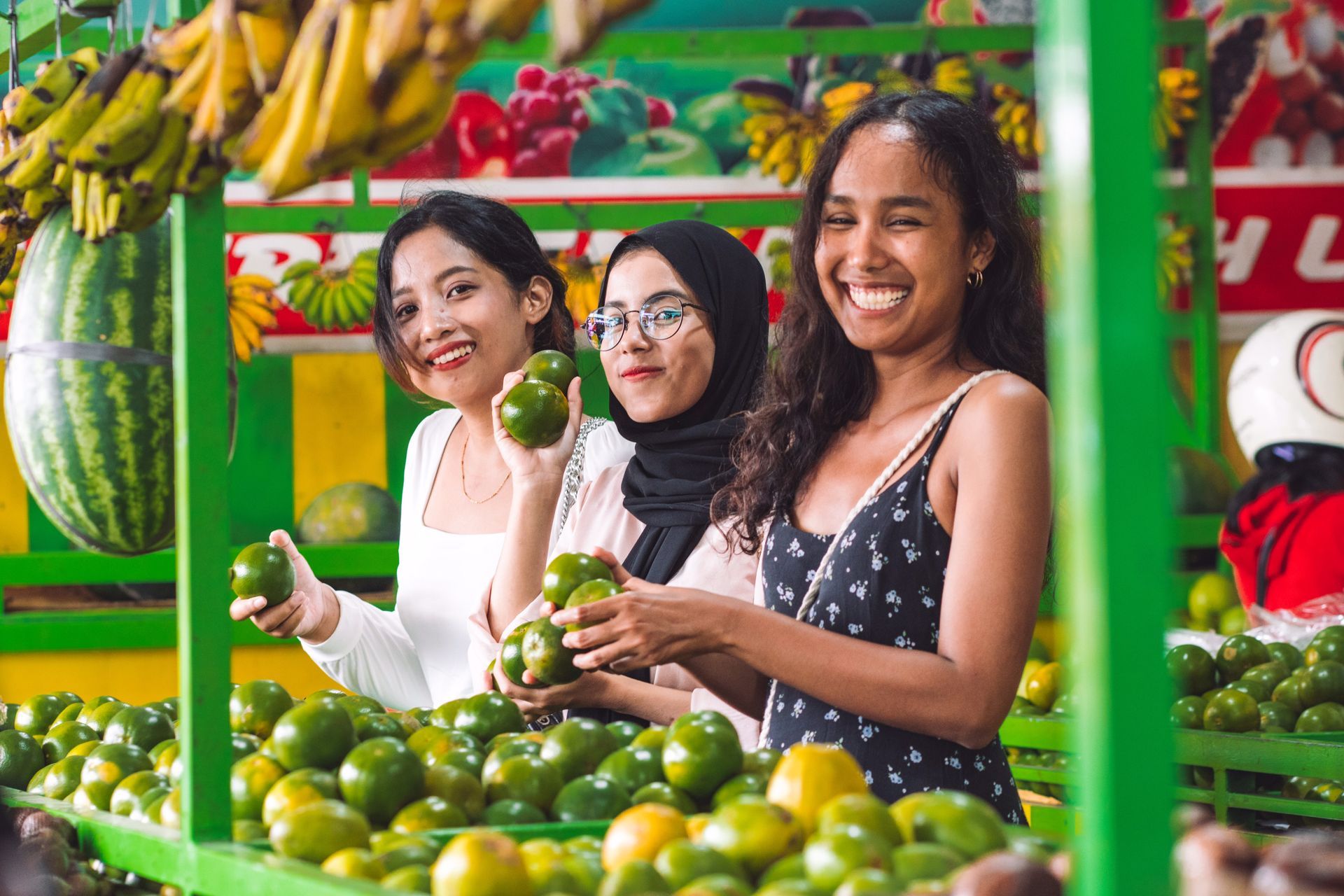 Tiga perempuan tersenyum, memetik alpukat di kios buah. Buah-buahan berwarna hijau dan kuning tampak di latar belakang.