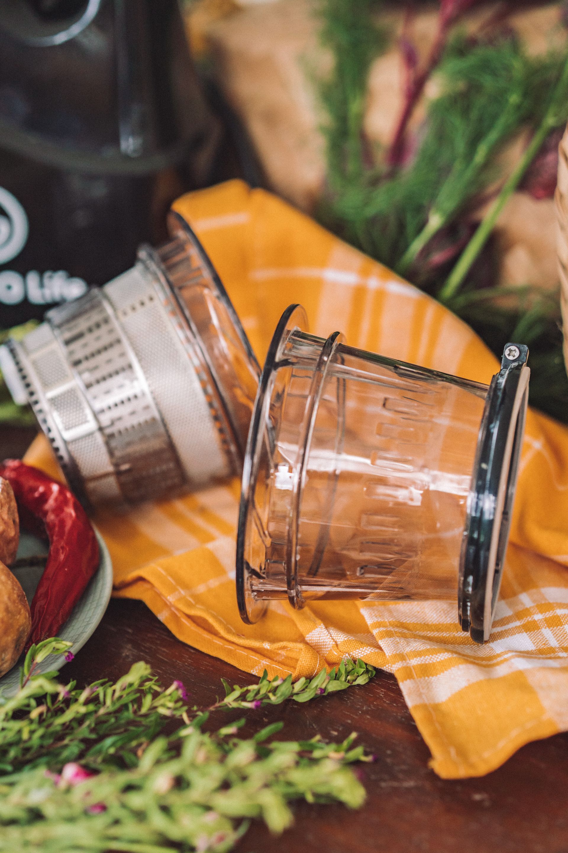 Dua saringan juicer di atas kain kotak-kotak kuning, dikelilingi oleh rempah-rempah dan sayuran.