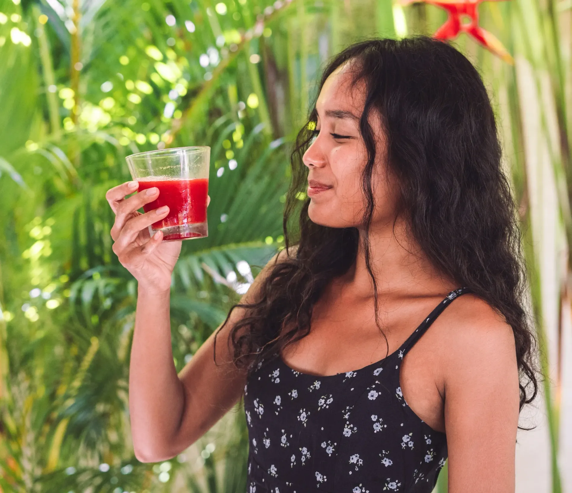 Wanita memegang segelas jus merah, tersenyum, dengan tanaman hijau di latar belakang.