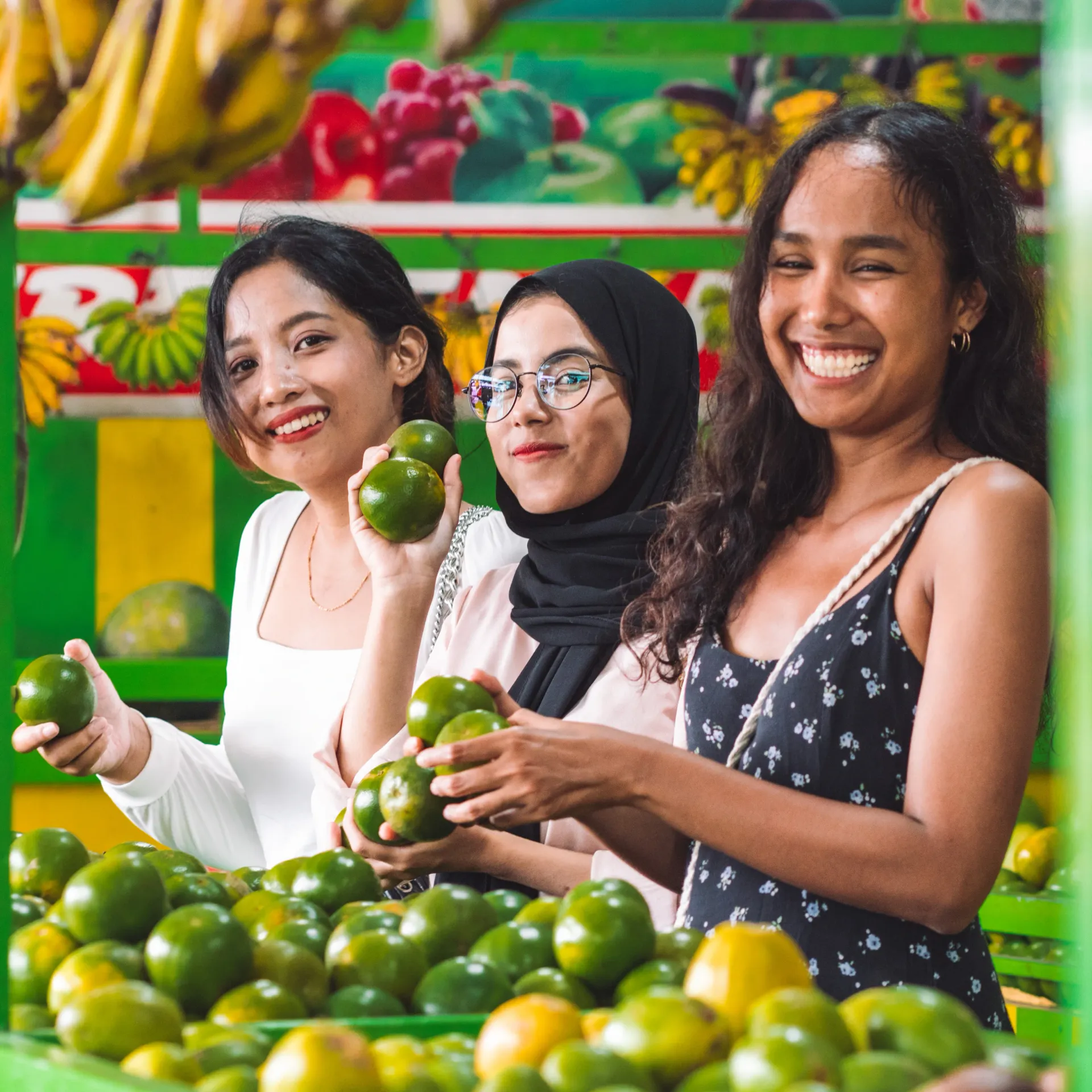 Tiga wanita tersenyum, memegang buah jeruk hijau di pasar.