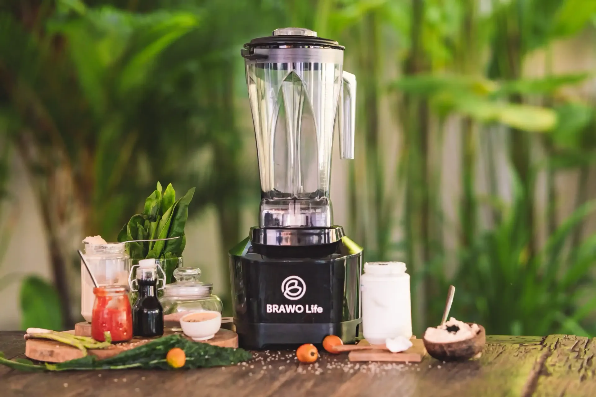 Blender hitam berisi bahan-bahan, diletakkan di atas meja kayu di luar ruangan dengan latar belakang pepohonan hijau.