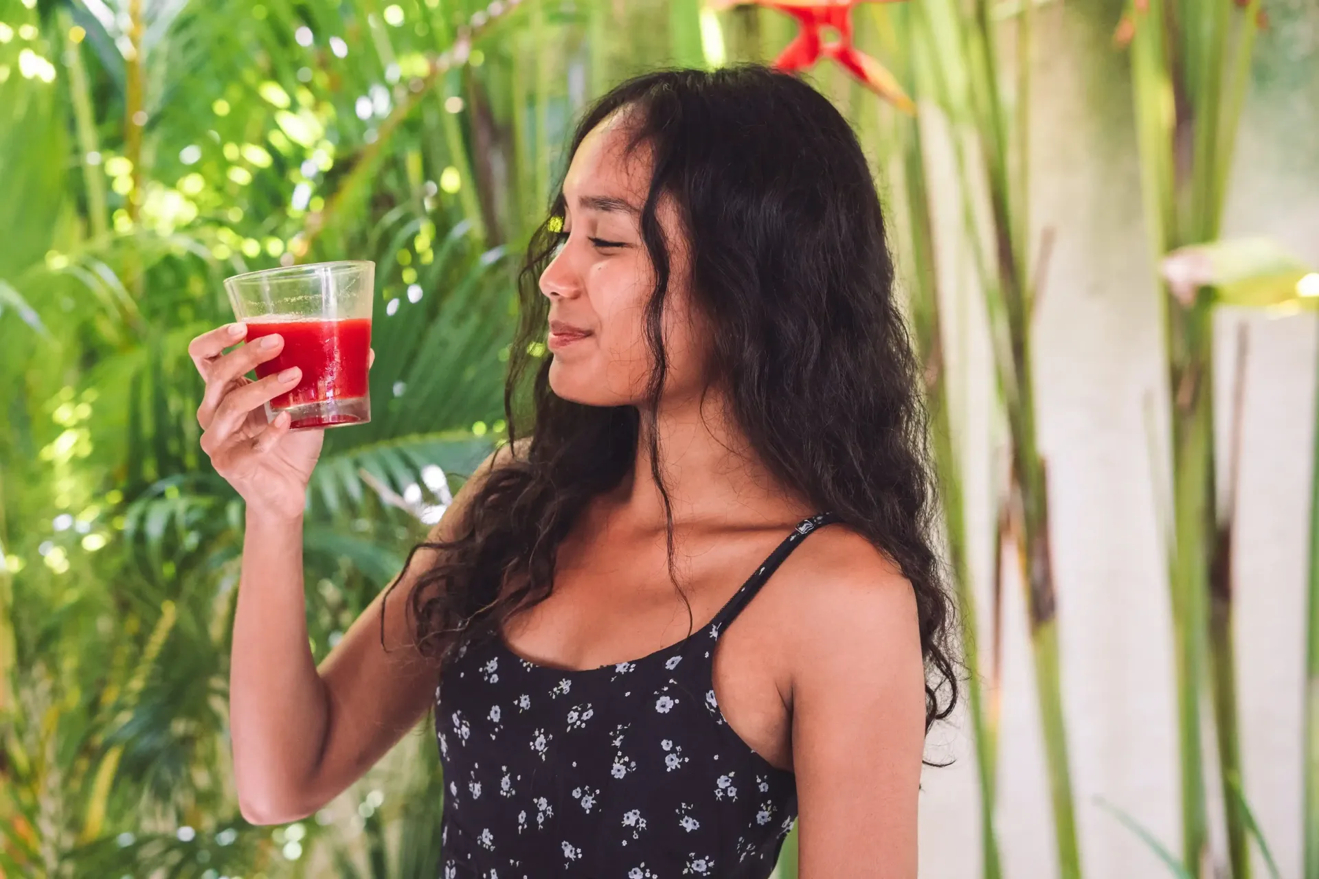 Wanita memegang dan mencium aroma jus merah, di luar ruangan dengan dedaunan hijau.