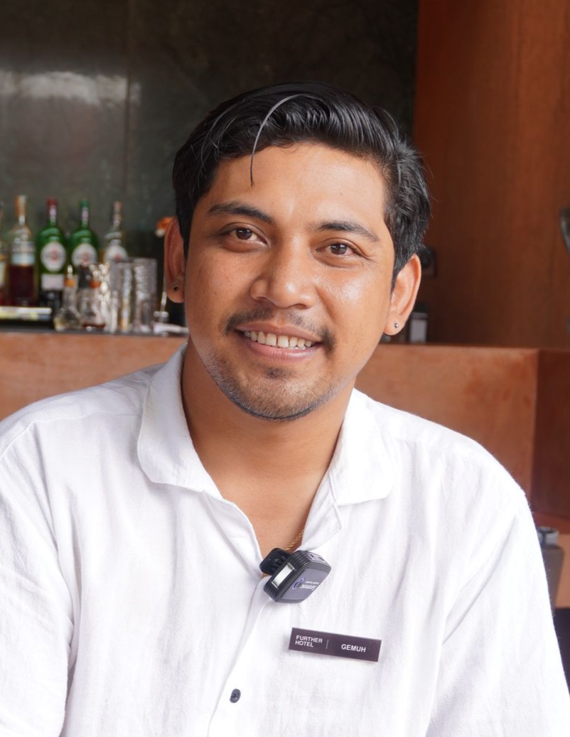 Seorang pria berambut gelap dan berjanggut tipis tersenyum, mengenakan kemeja putih; latar belakang botol-botol dan bar.