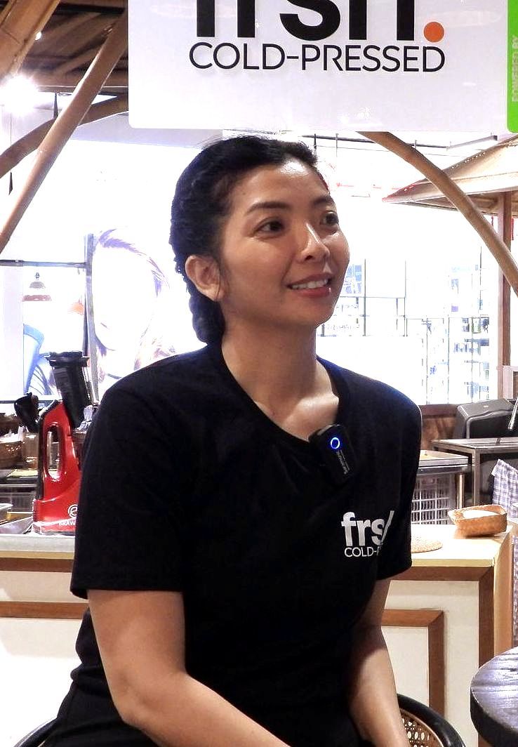 Seorang wanita berbaju hitam tersenyum, berdiri di dekat papan bertuliskan 