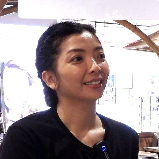 Wanita berambut hitam tersenyum, mengenakan kemeja hitam, di dalam ruangan.