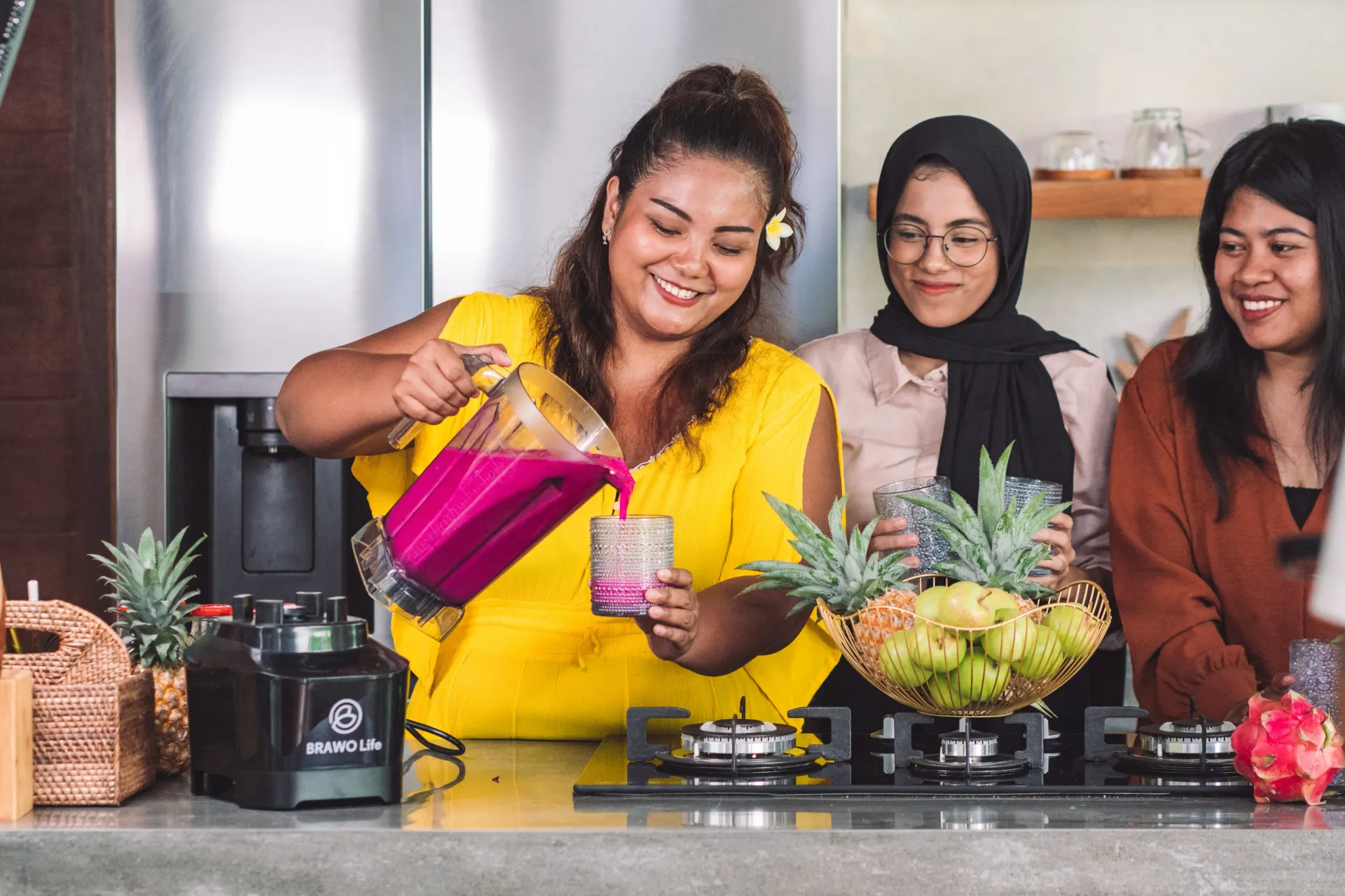 Tiga perempuan membuat smoothie; perempuan berbaju kuning menuangkan minuman merah muda. Dapur dengan buah-buahan.
