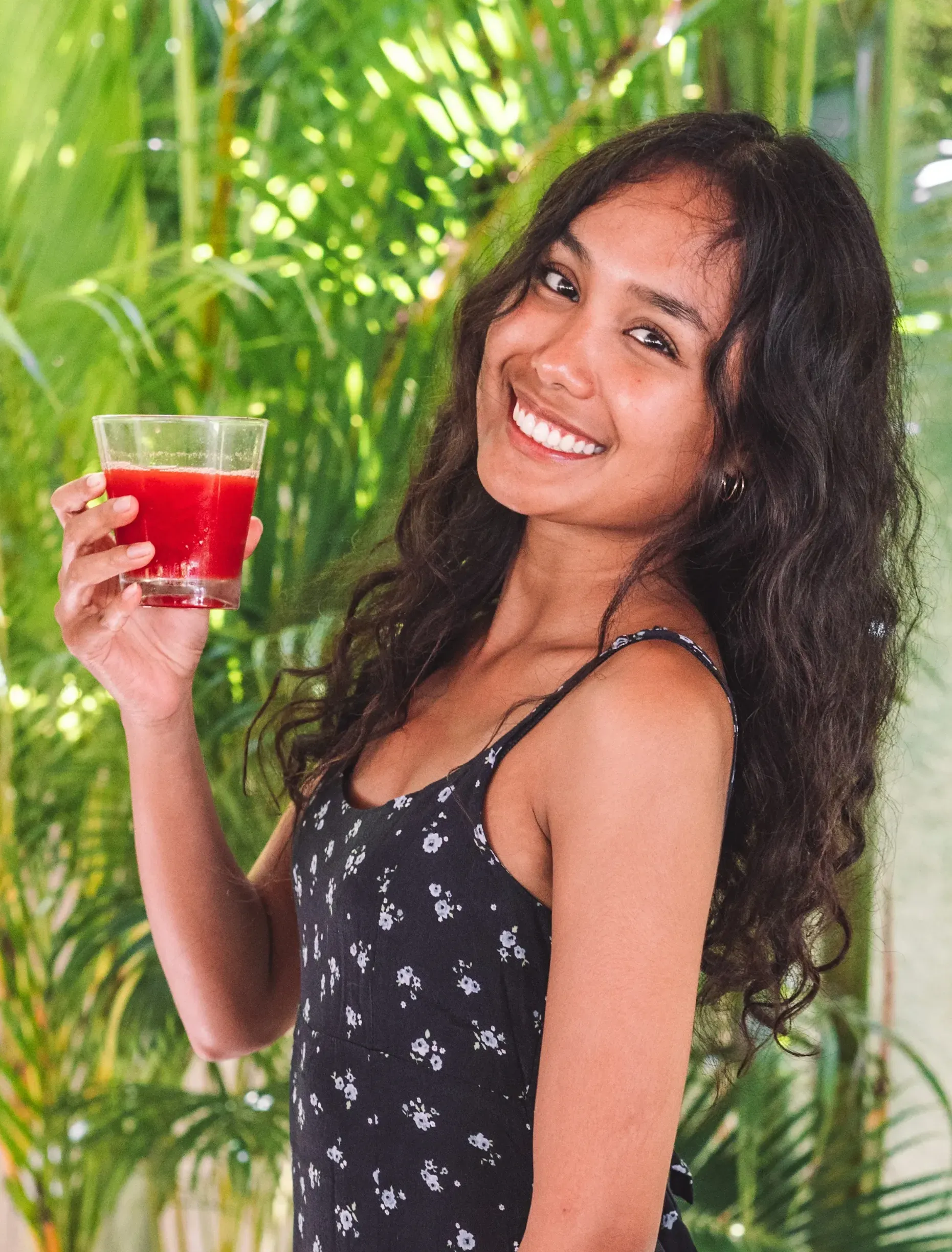 Seorang wanita berambut keriting tersenyum sambil memegang segelas jus merah di luar ruangan. Dedaunan hijau di latar belakang.