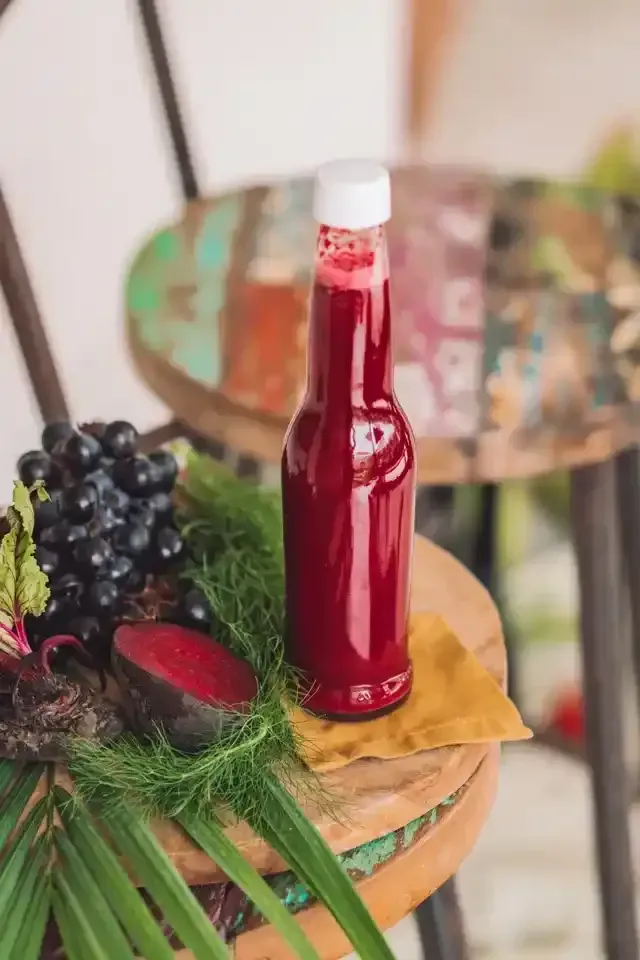 Sebotol jus merah di bangku pedesaan, dikelilingi anggur, bit, dan tanaman hijau.