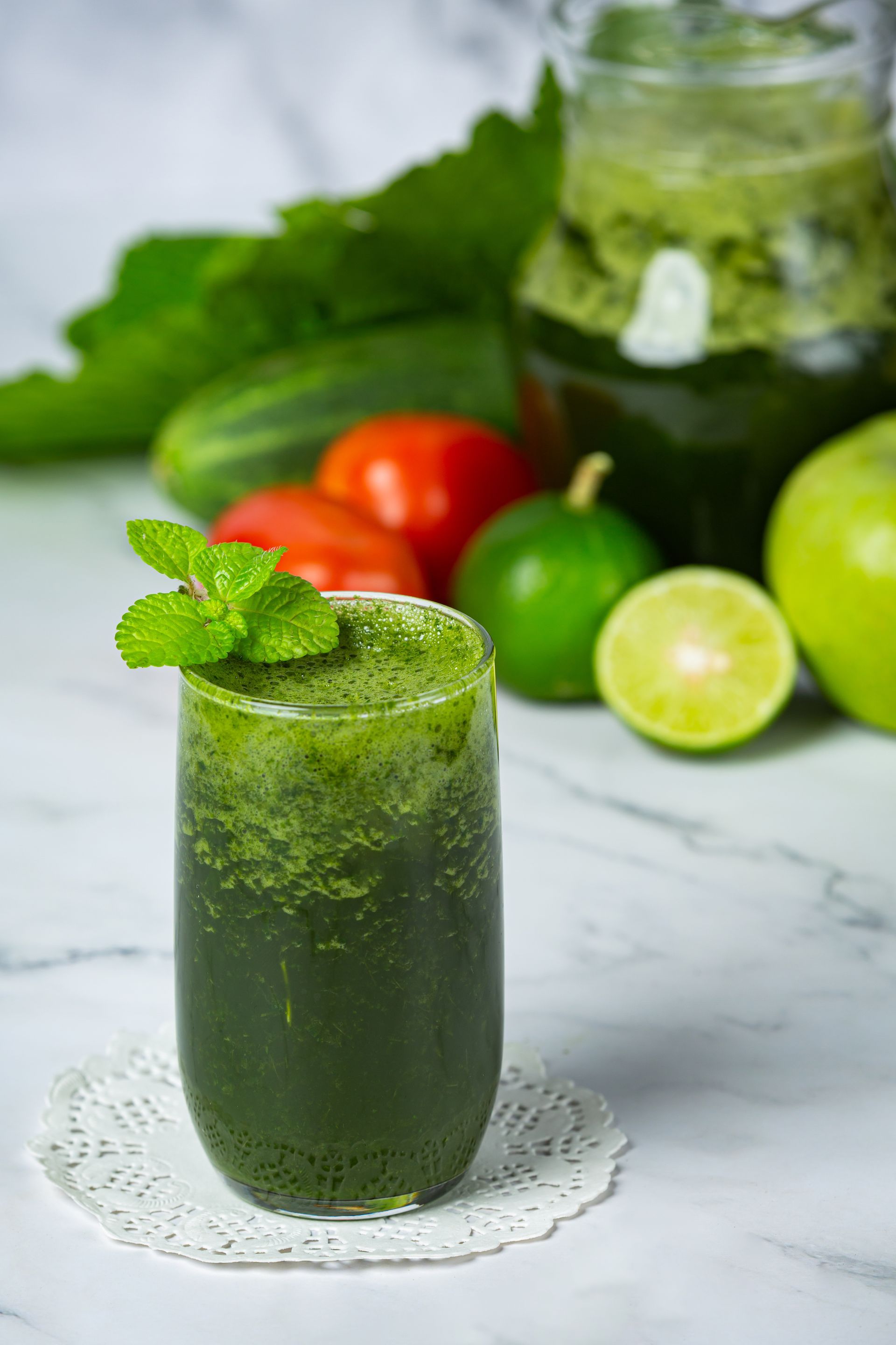 Jus hijau dalam gelas, dihiasi daun mint, dengan latar belakang sayuran dan buah.