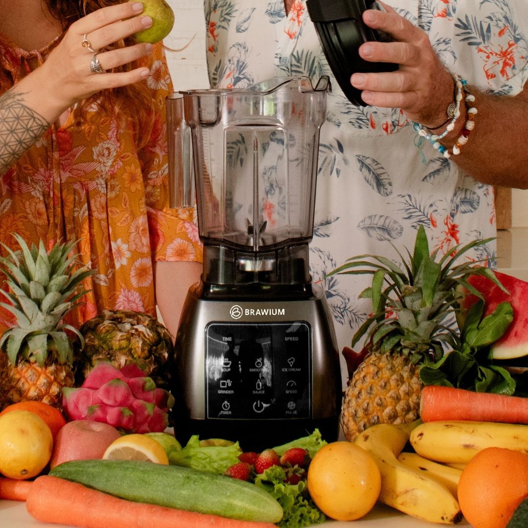 Blender dikelilingi buah, dengan dua orang sedang menyiapkan bahan-bahan.