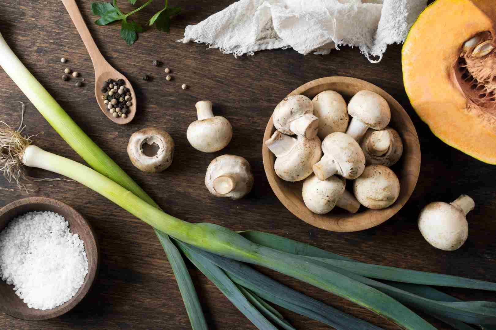 Jamur, daun bawang, labu, rempah-rempah, dan garam ditata di atas meja kayu.