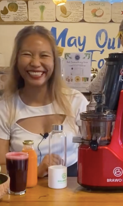 Seorang wanita tersenyum di samping juicer merah, memamerkan jus dan botol-botolnya. Suasana toko.